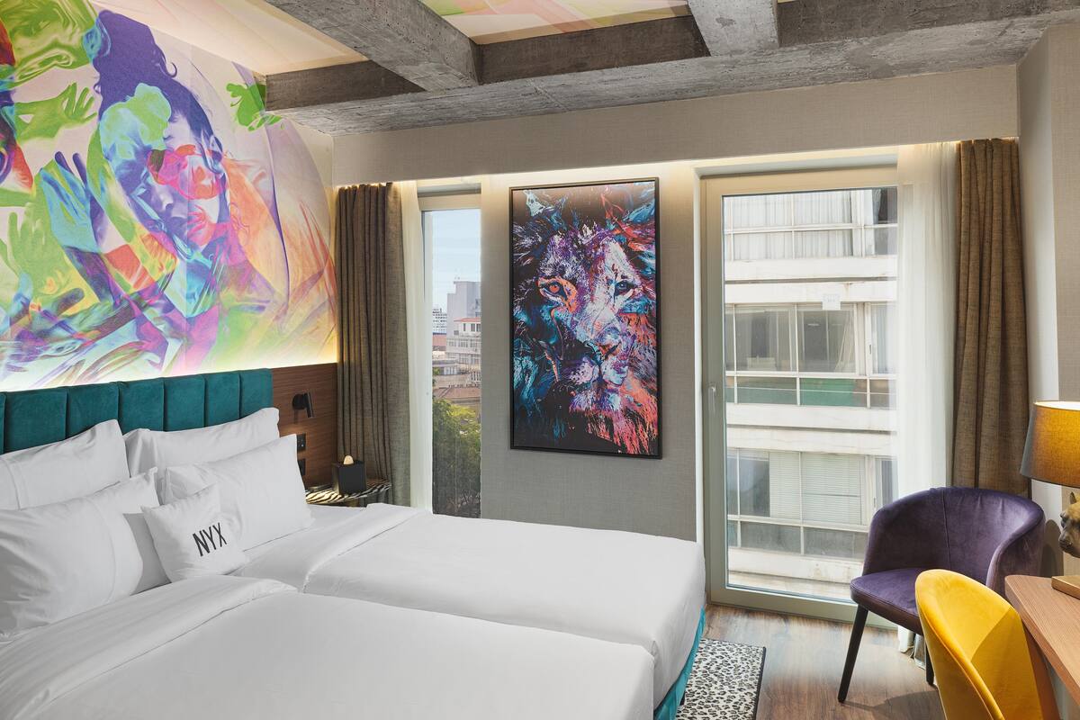 A hotel room with a large bed, a view of the city, and a colorful abstract painting on the wall.