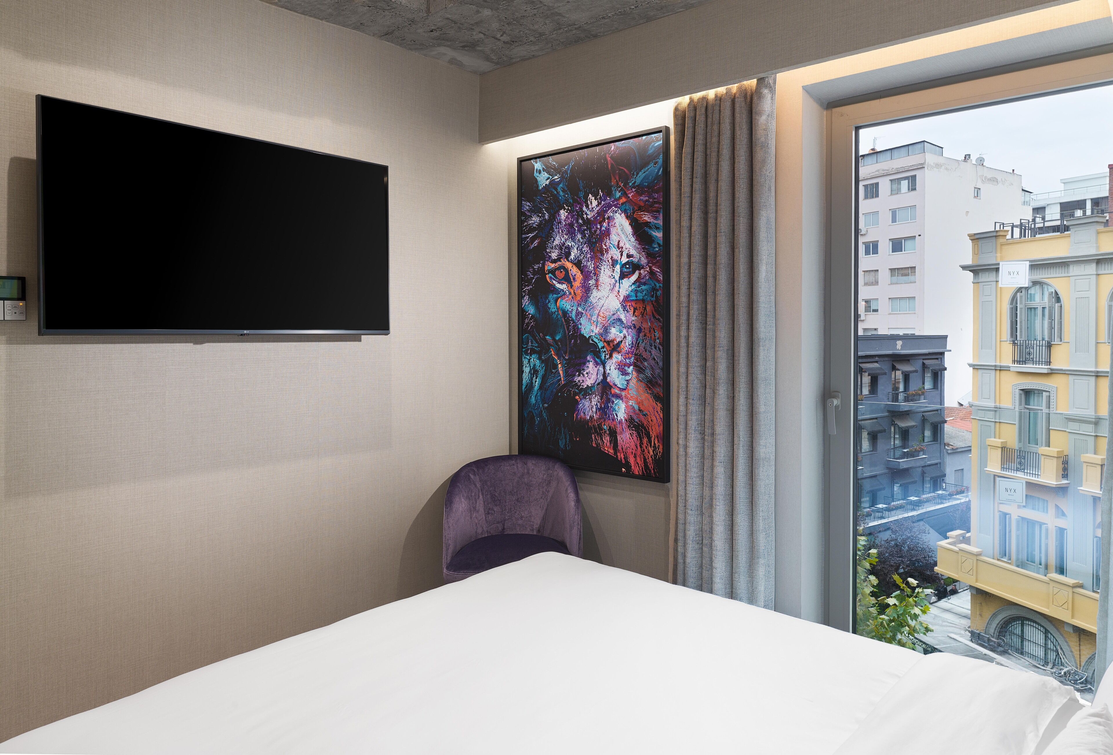 A hotel room with a bed, a flat-screen TV mounted on the wall, a large abstract painting, a window with curtains, and a view of buildings outside.