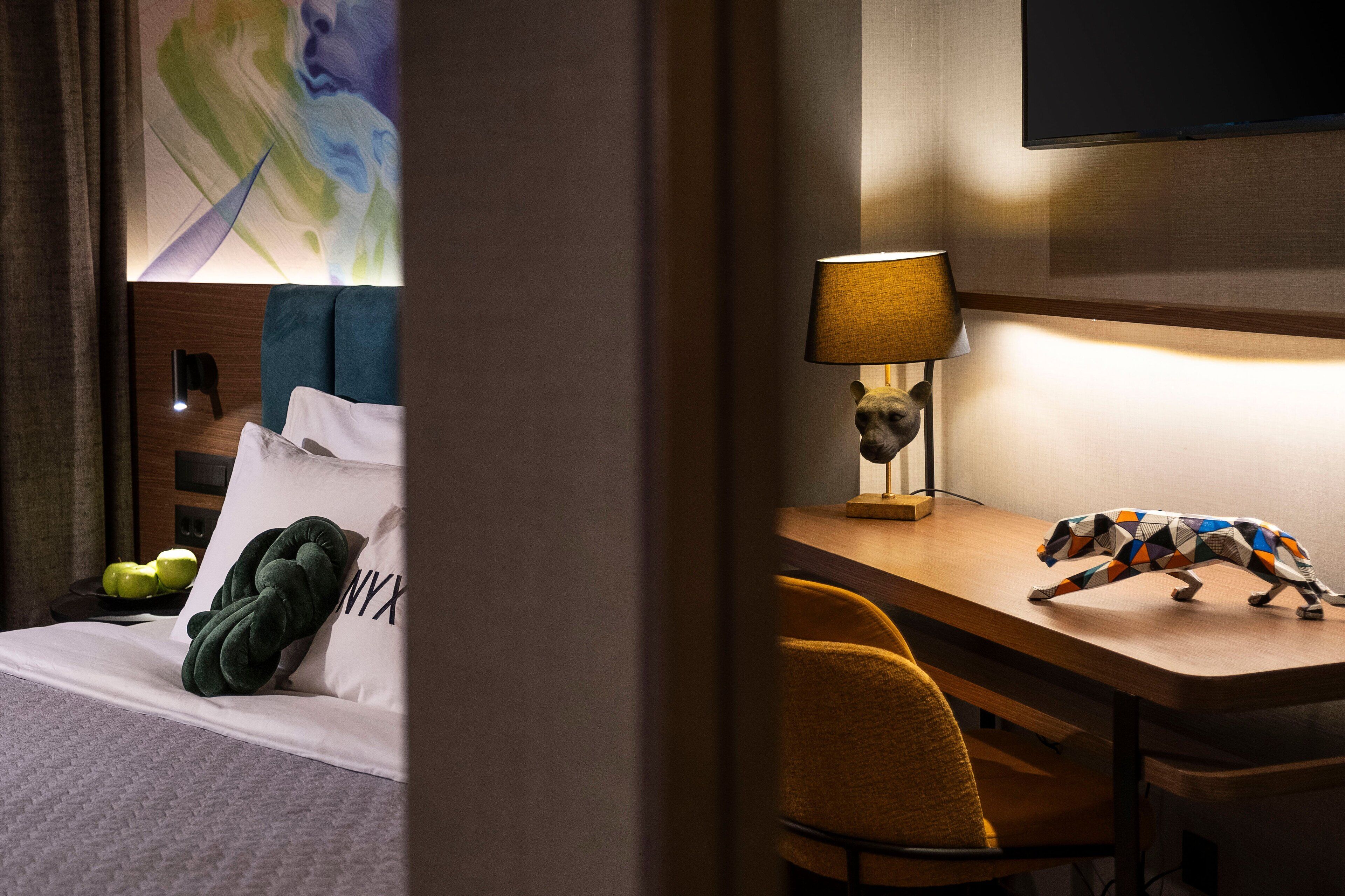 A hotel room with a bed, a desk with a lamp, a chair, and a wall-mounted TV.