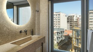 A modern bathroom with a circular mirror, a sink, and a large window offering a city view. - NYX Hotel Thessaloniki (Thessaloniki)