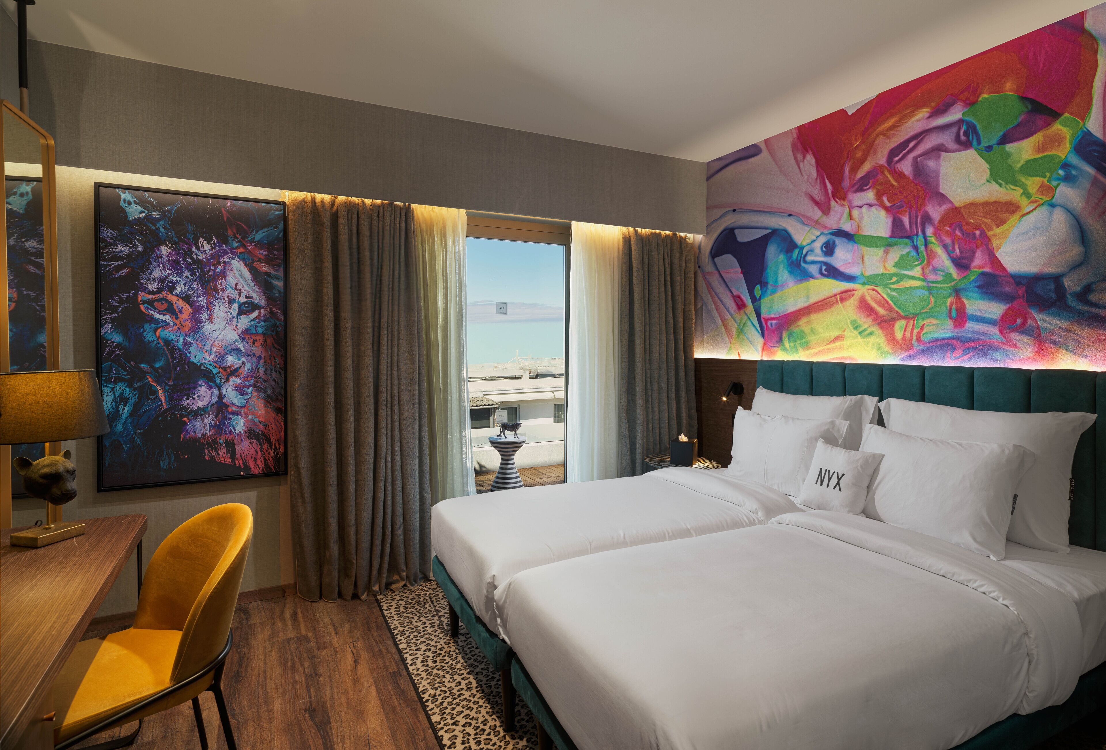 A hotel room with two beds, a large abstract painting, a desk with a lamp, and a view of the city.