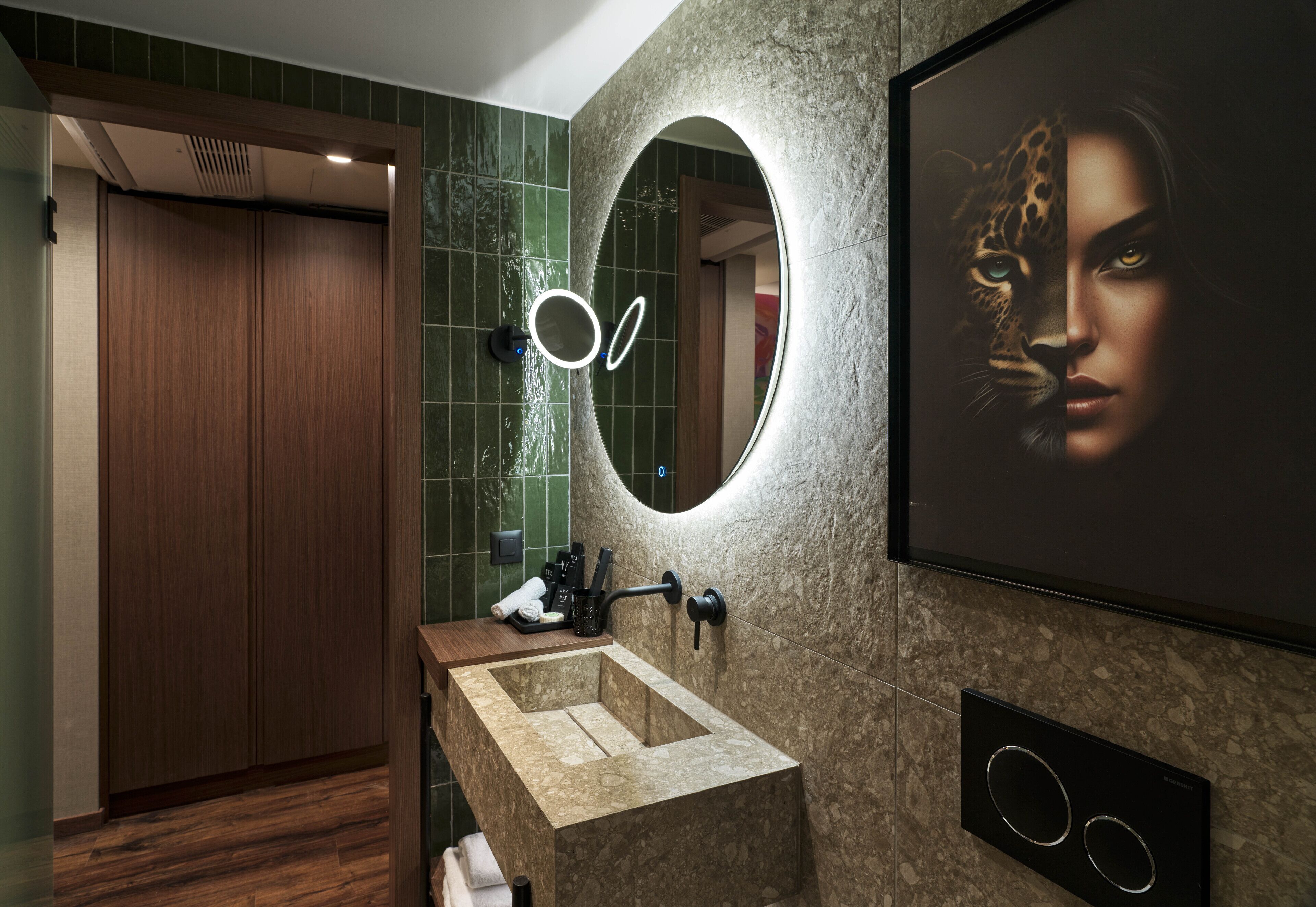 A modern bathroom with a wooden door, a mirror with circular light, and a large framed picture on the wall.