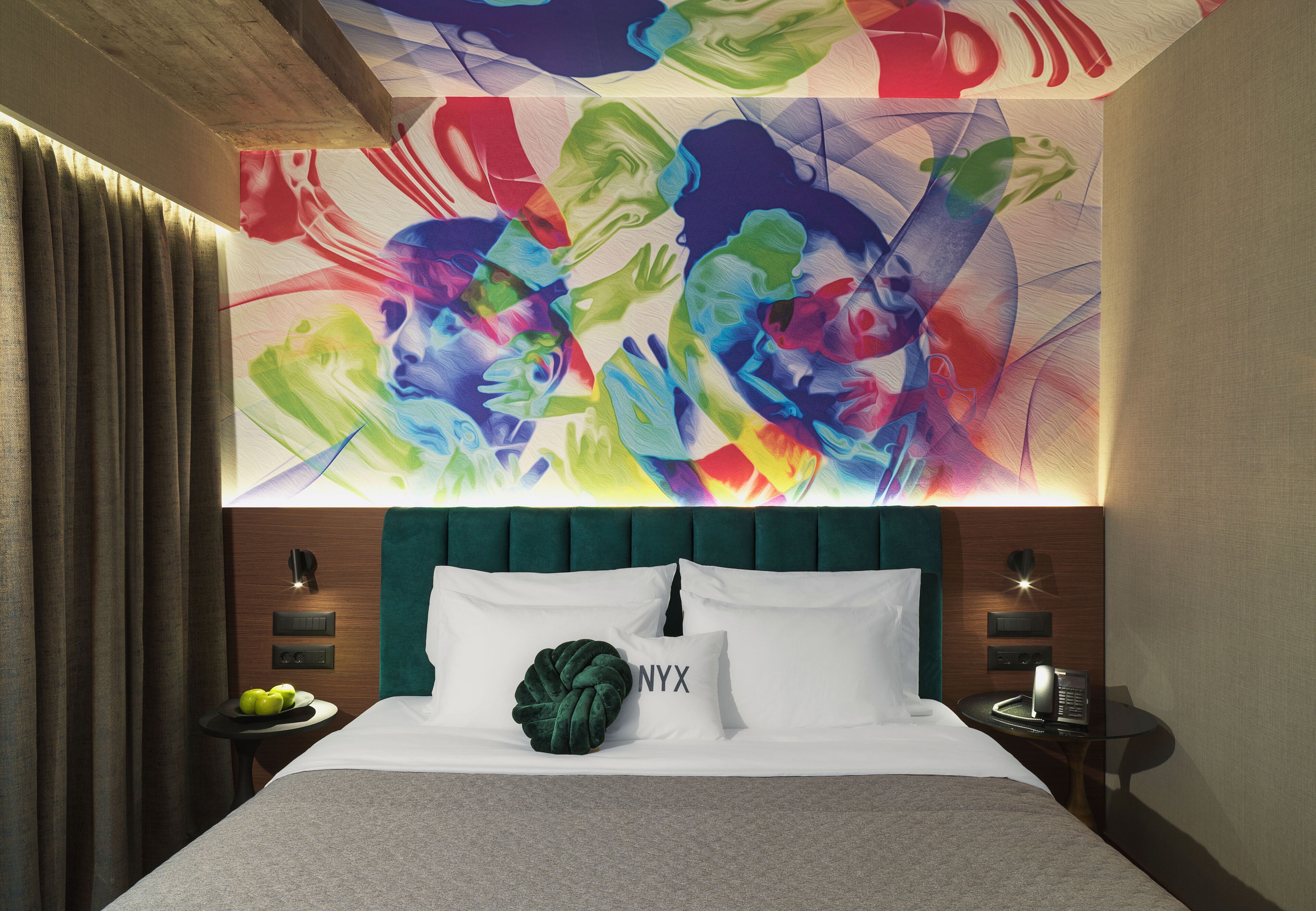 Una habitación de hotel con una cama grande, un cabecero tapizado en verde, un mural en la pared, una mesita de noche con un teléfono y una pequeña mesa con manzanas.