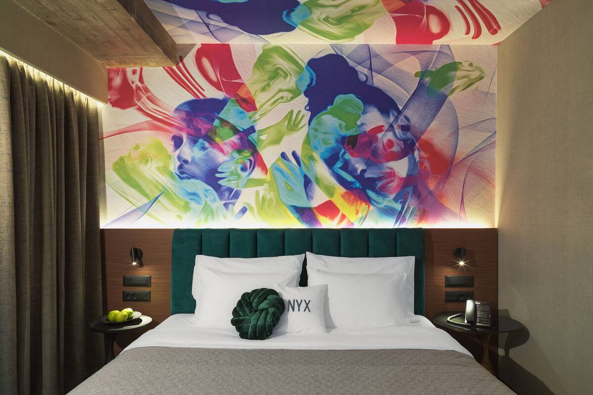 A hotel room with a large bed, a green upholstered headboard, a wall mural, a bedside table with a phone, and a small table with apples.
