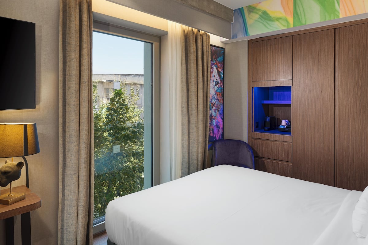 A hotel room with a bed, a desk with a lamp, a chair, a TV, and a window with curtains.