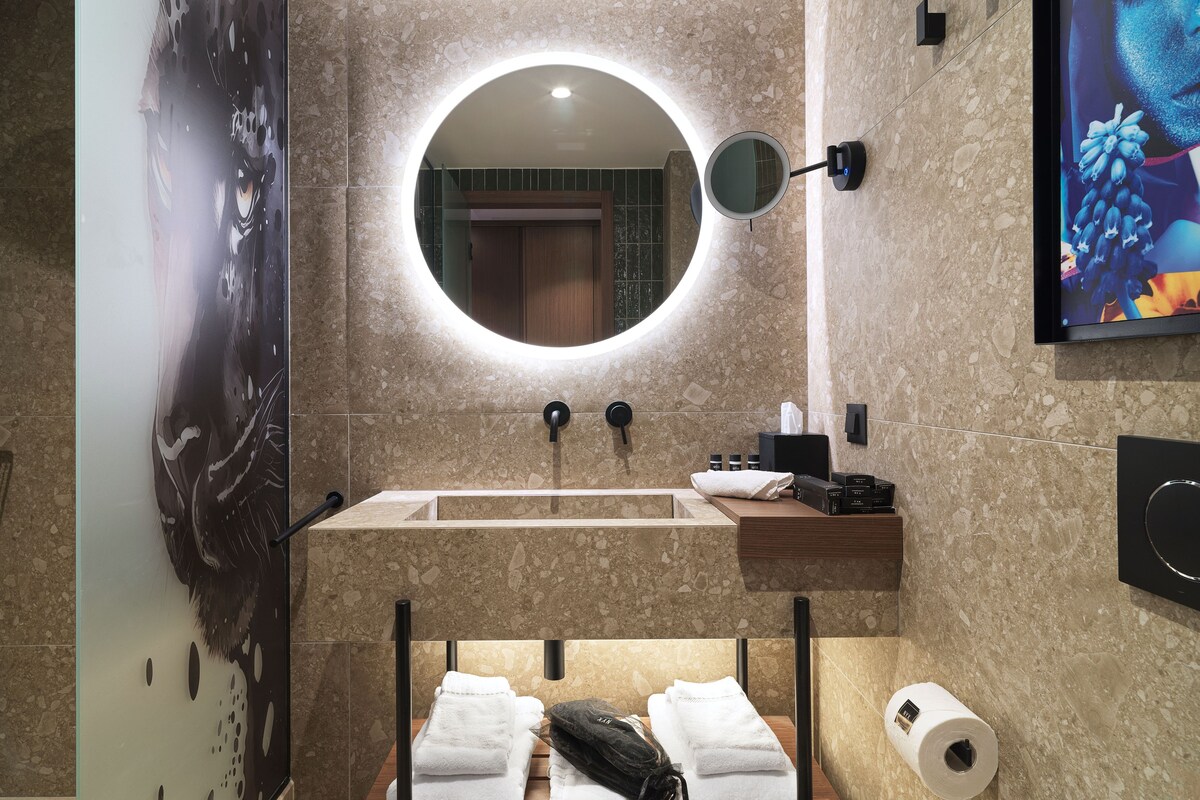 A bathroom with a round mirror, sink, towel rack, and toilet paper dispenser.
