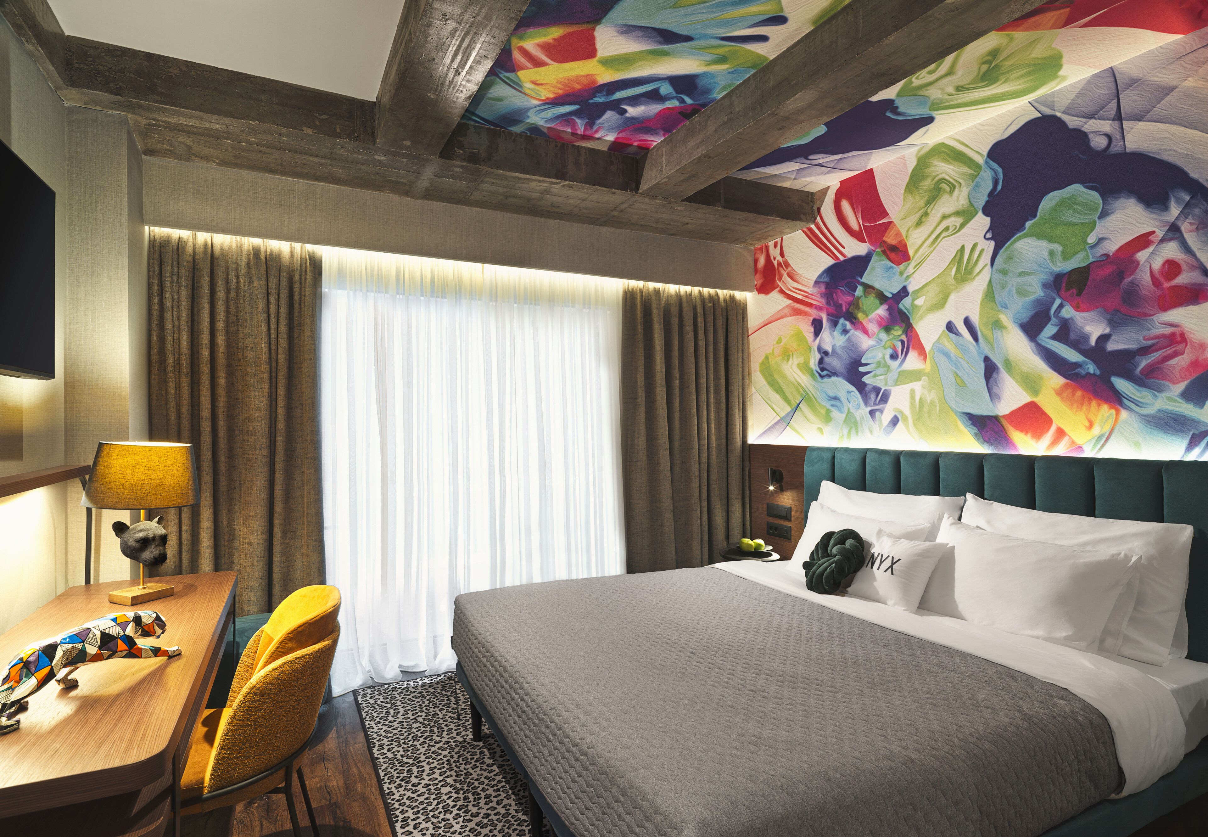 Una habitación de hotel con cama, escritorio con lámpara, silla, televisor y un mural colorido en la pared.