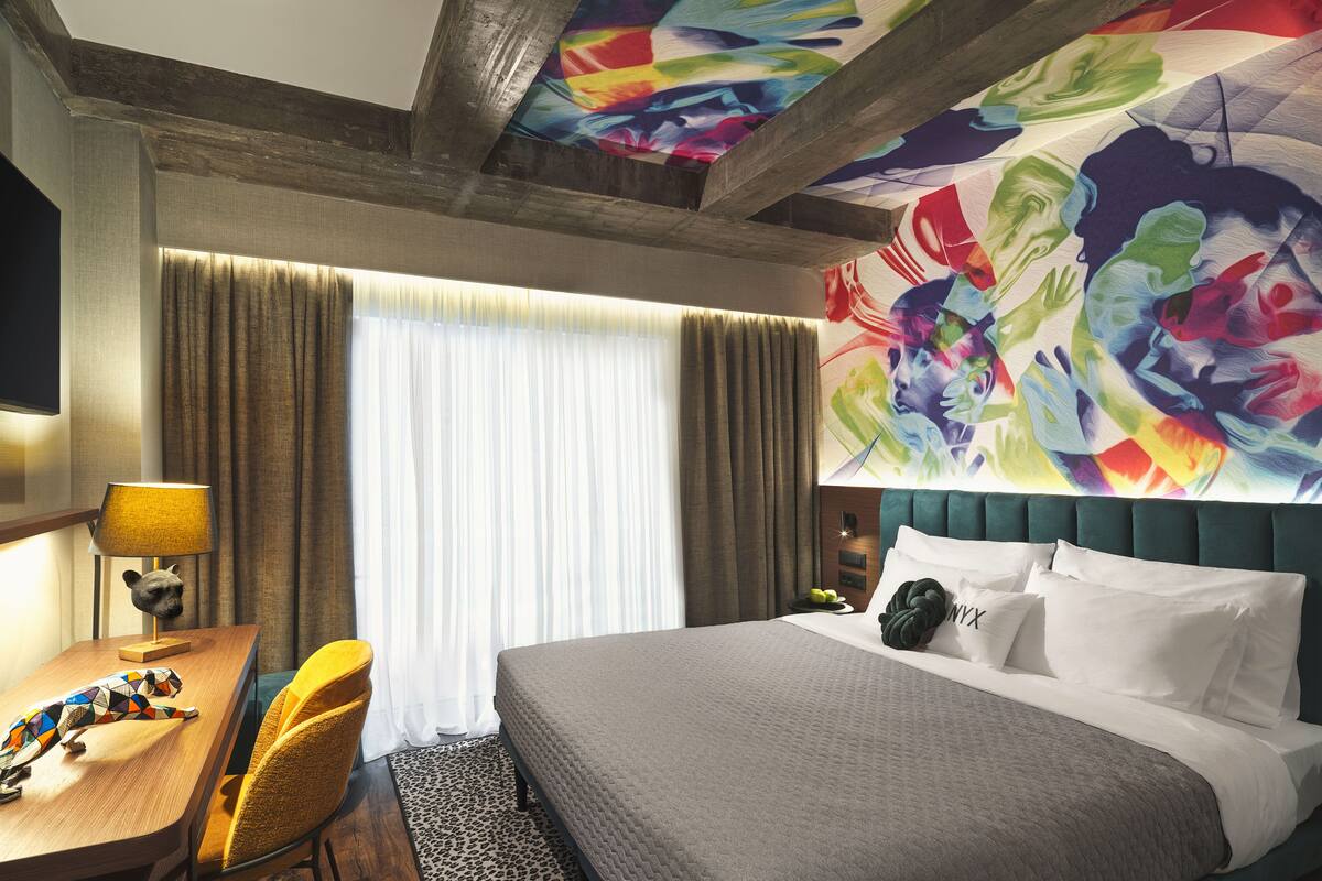 A hotel room with a bed, a desk with a lamp, a chair, a TV, and a colorful mural on the wall.