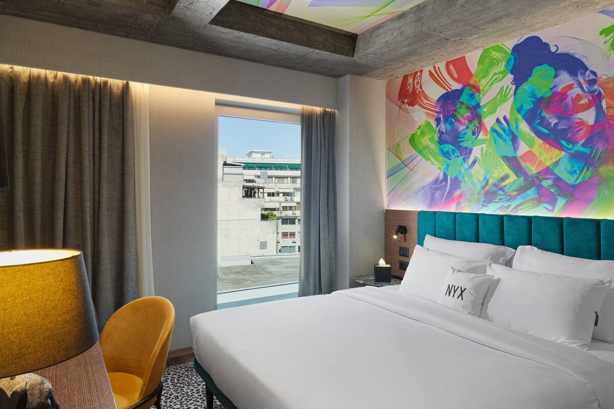 A hotel room with a large bed, a view of the city, and a colorful mural on the wall.