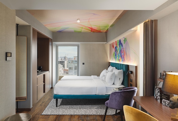 Habitación de hotel con cama, escritorio, silla, ventana con vista a la ciudad y un cuadro abstracto colorido en la pared.