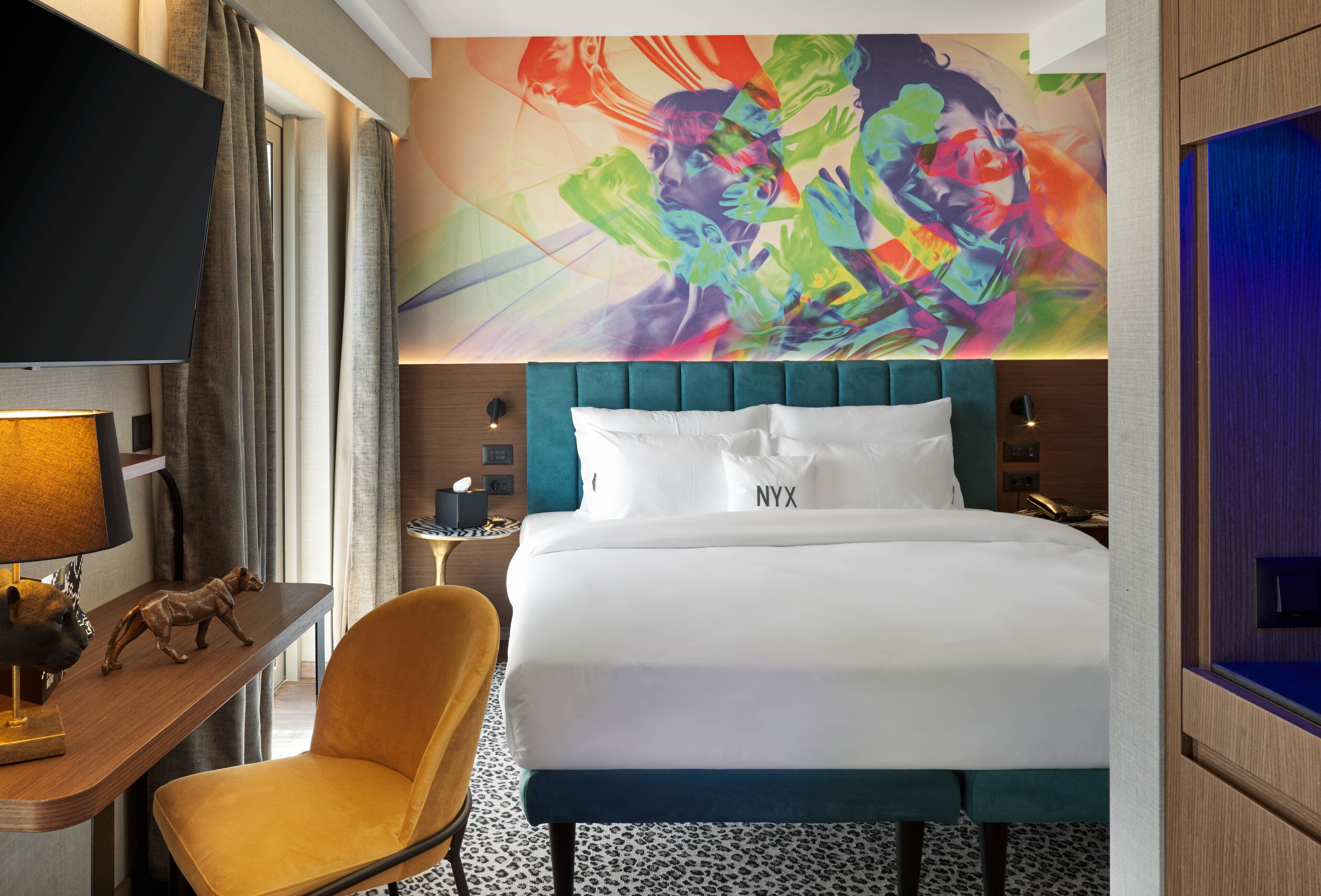 Habitación de hotel con una cama grande, televisión, escritorio con lámpara, silla y un cuadro abstracto colorido en la pared.