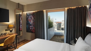 A hotel room with a large bed, a desk with a lamp, a chair, a painting, and a view of the city through the window. - NYX Hotel Thessaloniki (Thessaloniki)