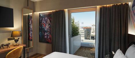 Une chambre d’hôtel avec un grand lit, un bureau avec une lampe, une chaise, un tableau et une vue sur la ville par la fenêtre.