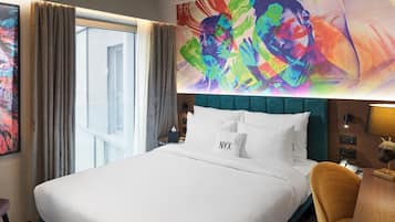 A hotel room with a large bed, a teal headboard, a colorful abstract mural, a desk with a lamp, and a leopard print rug.