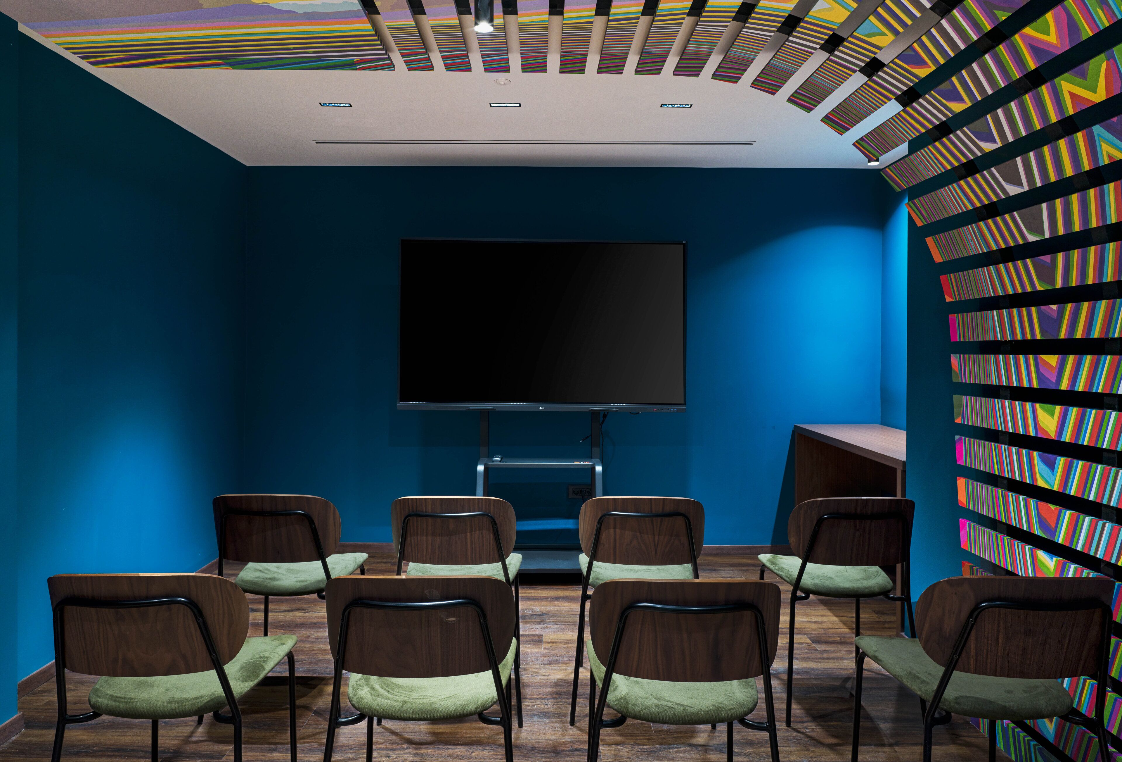 A conference room with a large flat-screen TV, six wooden chairs with green cushions, and a colorful striped wall on the right.