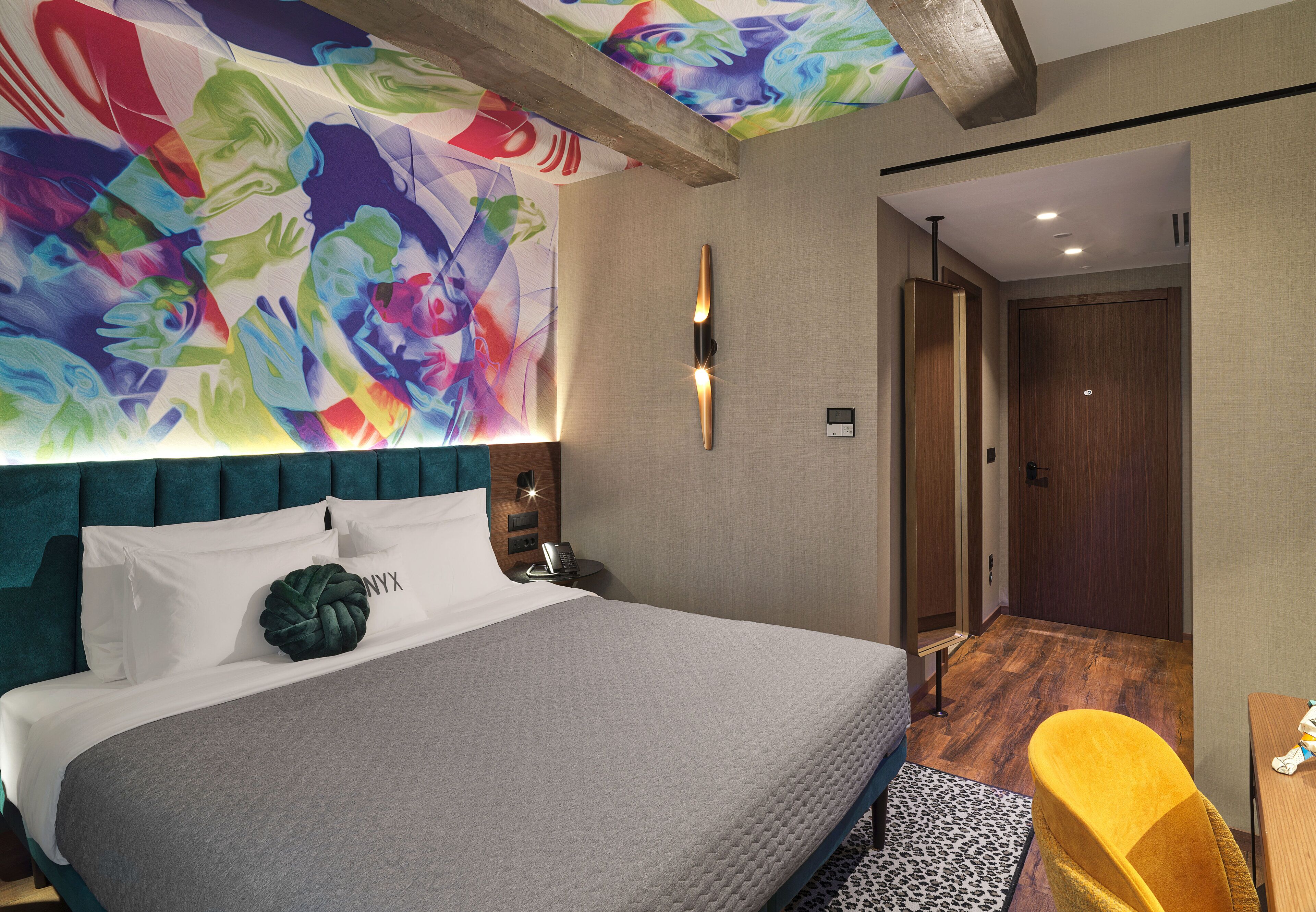 A modern hotel room with a large bed, a vibrant abstract mural on the wall, a wooden door, and a yellow chair.