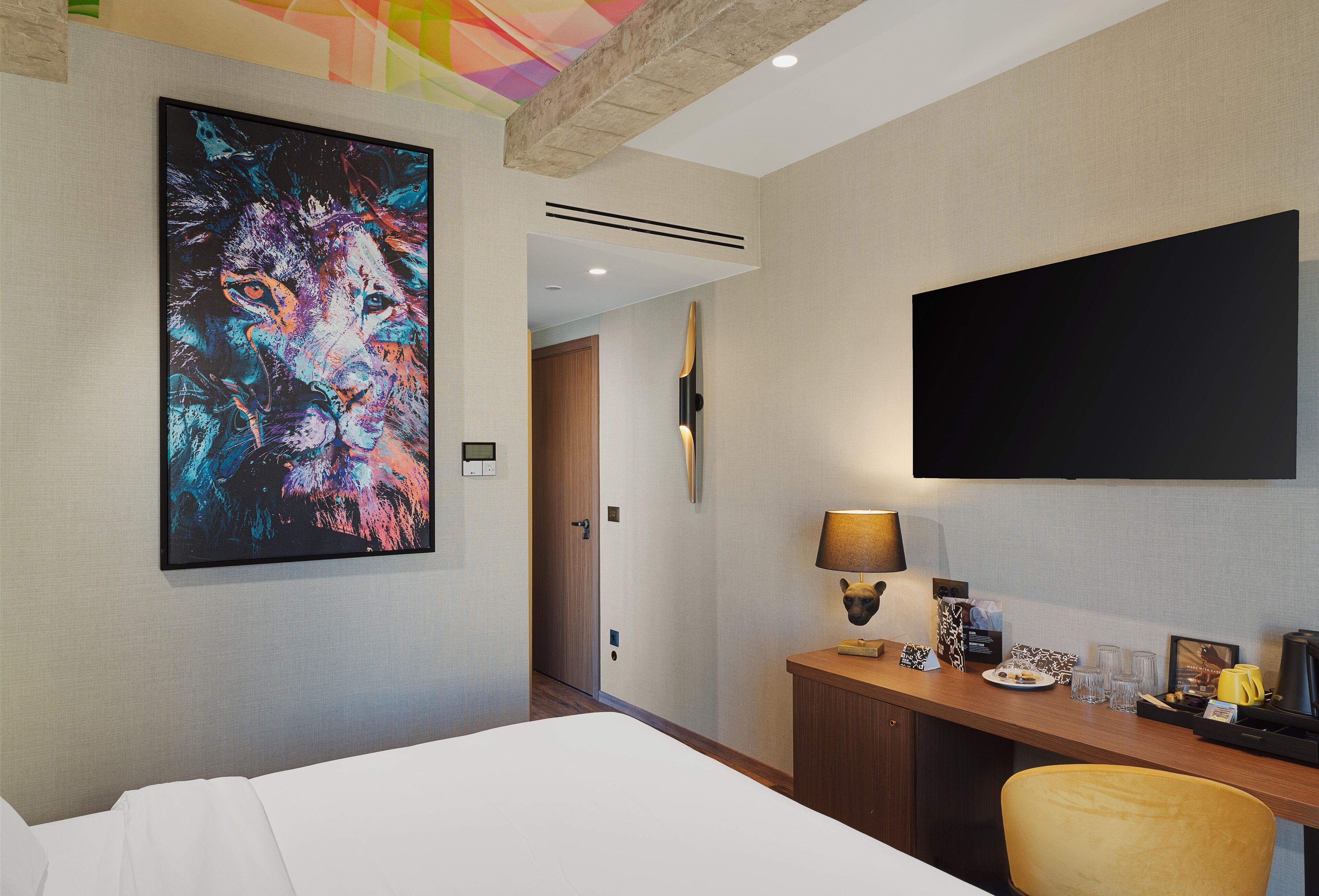 A hotel room with a bed, a desk with a lamp, a flat-screen TV, and a large abstract painting on the wall.