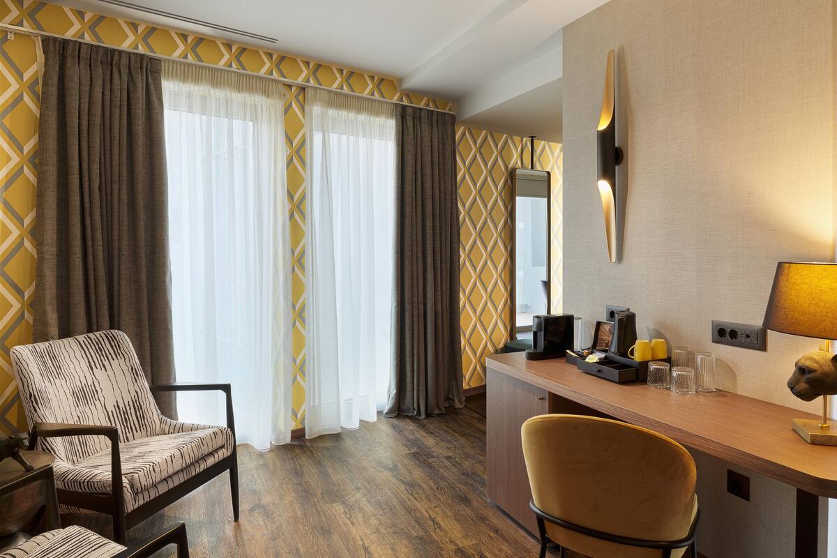 A hotel room with a wooden desk, two chairs, a sofa, a large window with curtains, a wall-mounted lamp, and a mirror.