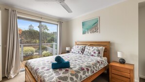 Un dormitorio con un ventanal grande, una cama con ropa de cama estampada, un cabecero de madera, una mesita de noche y un ventilador de techo.