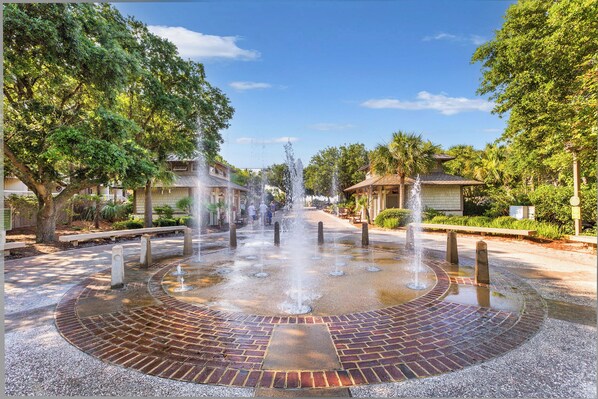 A circular brick fountain with water jets in a landscaped area with trees and buildings.