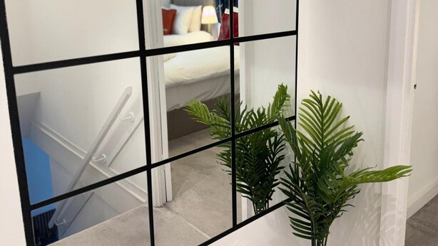 Un couloir avec un mur miroitant qui reflète une chambre à coucher avec un lit et une table de chevet. On aperçoit un escalier avec une rampe dans le reflet.