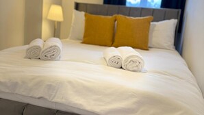 Un lit bien fait, avec du linge de lit blanc et des serviettes enroulées, une tête de lit grise et un coussin de couleur moutarde.