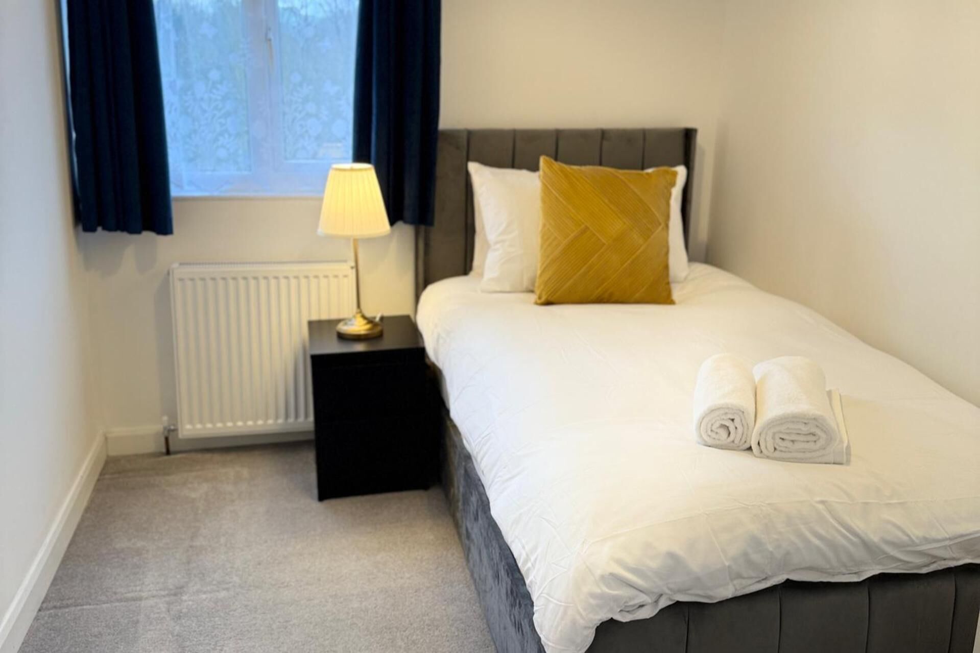 A neatly made bed with white linens and a rolled towel, a bedside lamp, and a window with curtains.