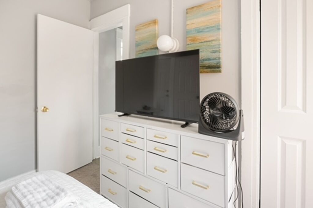 Une chambre avec une commode blanche, un téléviseur posé dessus et un ventilateur sur pied.