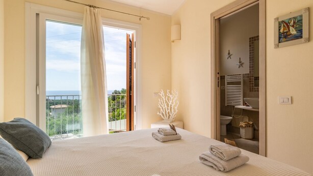 A bedroom with a bed, a bathroom visible through an open door, and a balcony with a view.