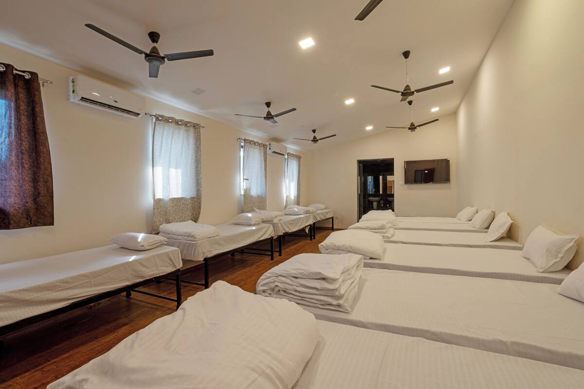 Habitación de hotel con varias camas individuales dispuestas en filas, ventiladores de techo y un equipo de aire acondicionado.