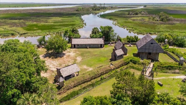 An aerial view of a rural property with several wooden buildings, a fenced area, and a water body.
