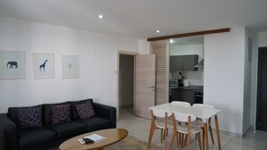 Una sala de estar moderna con un sofá negro, una mesa de centro de vidrio y un comedor con sillas blancas. Hay impresiones enmarcadas de animales en la pared y se ve una cocina a través de una puerta abierta.