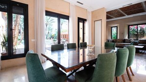 A spacious dining area with a large glass table and green upholstered chairs, large windows with curtains, and a view of palm trees and a patio outside. - Luxury villa on 2200m2, restaurants and supermarket 5 minutes from the center, 6km from the town center. (Marrakech)