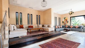A spacious living room with a large sectional sofa, a coffee table, and a colorful rug. There are large windows and a view of greenery outside. - Luxury villa on 2200m2, restaurants and supermarket 5 minutes from the center, 6km from the town center. (Marrakech)