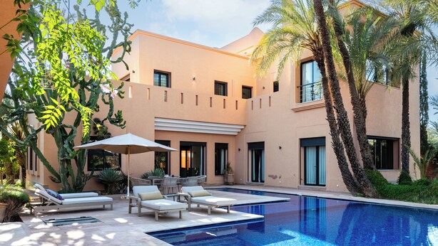 A modern pool area with lounge chairs and a large umbrella, surrounded by palm trees and a building with a beige facade. - Luxury villa on 2200m2, restaurants and supermarket 5 minutes from the center, 6km from the town center. (Marrakech)