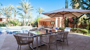 An outdoor dining area with a glass table, wicker chairs, and a large umbrella. - Luxury villa on 2200m2, restaurants and supermarket 5 minutes from the center, 6km from the town center. (Marrakech)
