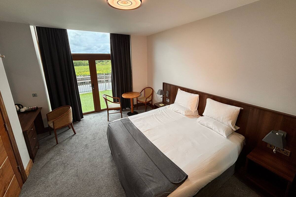 Habitación de hotel con una cama grande, una mesita pequeña, dos sillas, una mesita de noche y vistas a un área verde a través de una ventana.