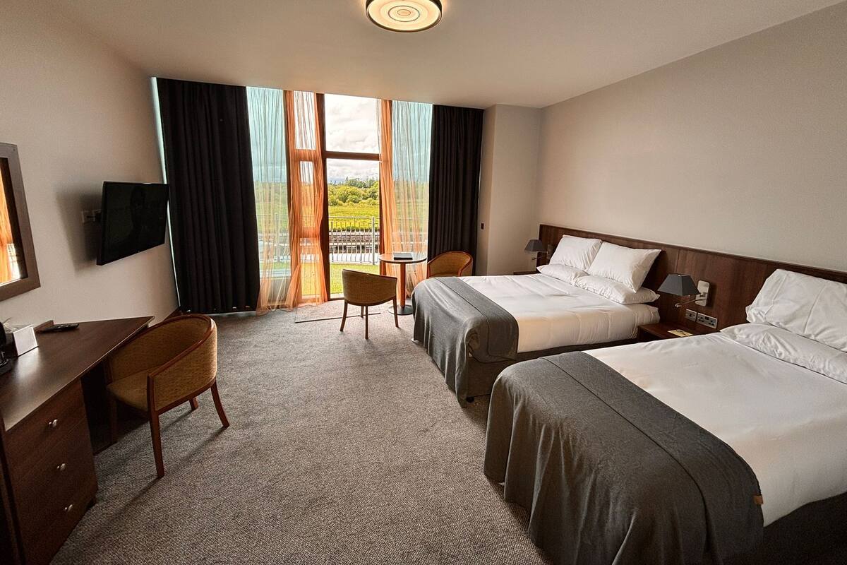 Habitación de hotel con dos camas, un escritorio, una silla, televisión y vista a un paisaje verde a través de amplios ventanales.