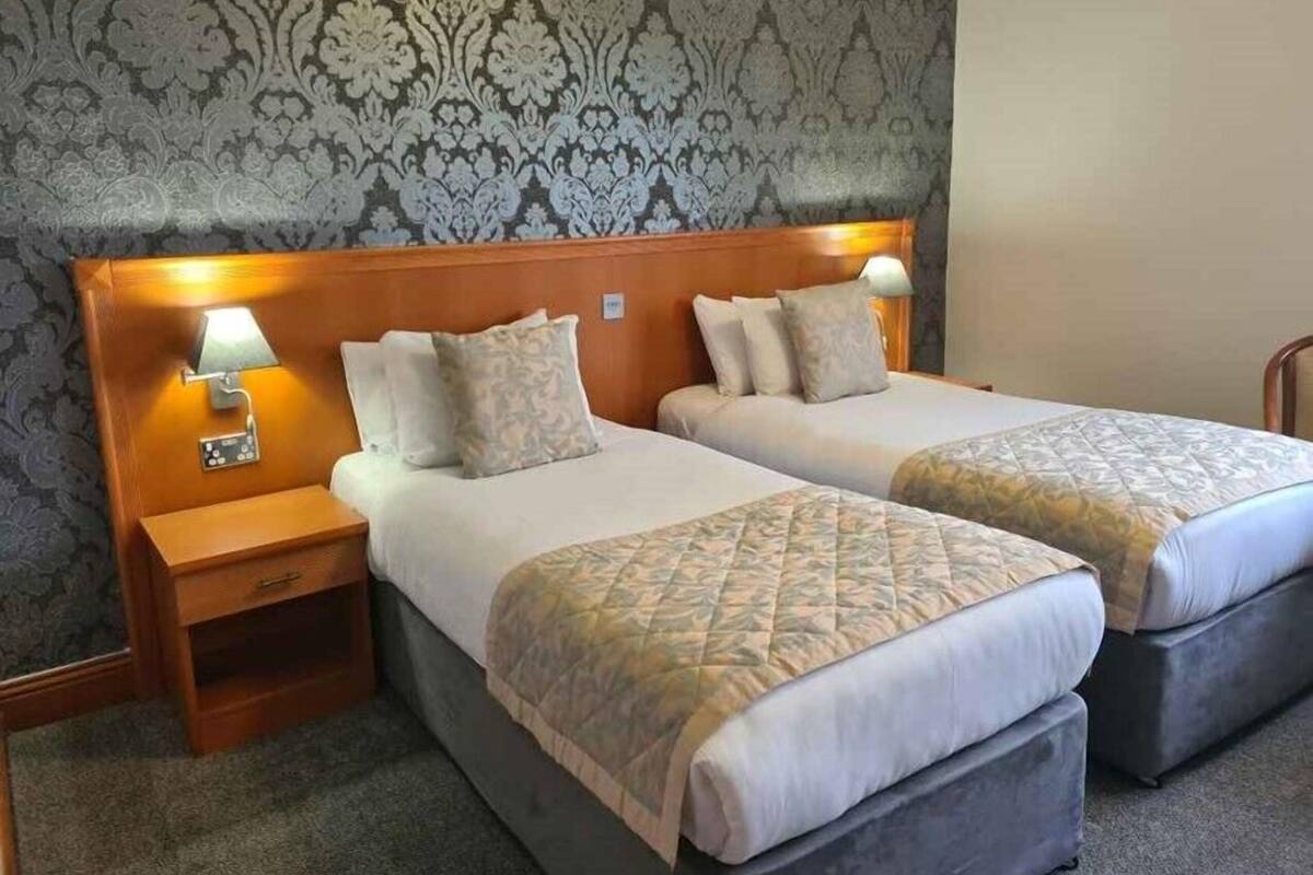 Una habitación de hotel con dos camas, una mesita de noche con lámpara y un papel tapiz estampado.