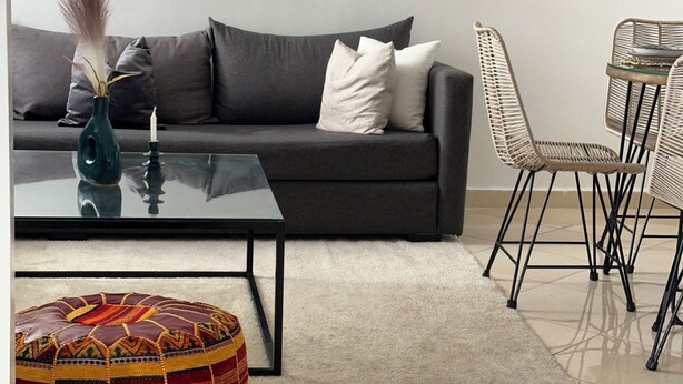 A modern living room with a dark grey sofa, a glass coffee table, a colorful pouf, and a dining area with wicker chairs.