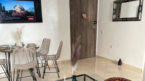 A modern living room with a glass coffee table, wicker chairs, a TV mounted on the wall, and a wooden door.