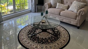 A living room with a beige sofa, a glass coffee table with candles, and a view of greenery through large windows.