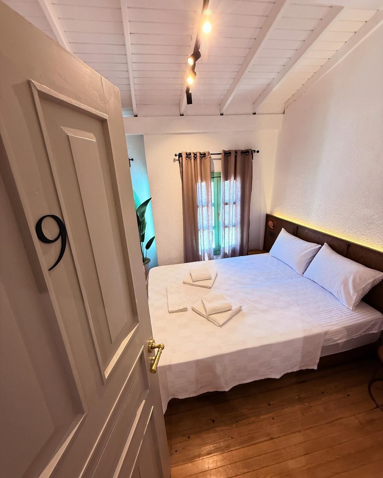 A hotel room with a large bed, a wooden floor, a door with the number 9, and a ceiling with exposed beams and lighting.