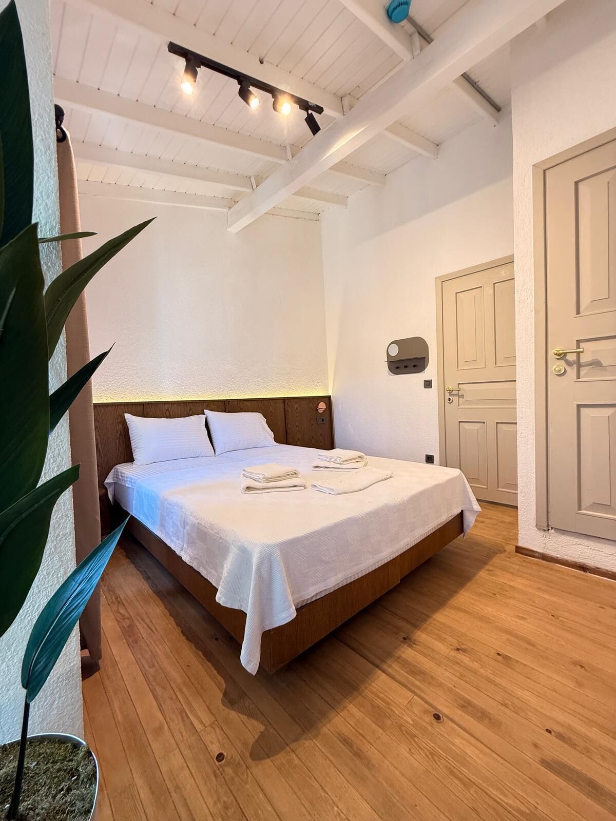 A neatly made bed in a room with wooden flooring, a white headboard, and a ceiling with exposed beams and lighting fixtures.