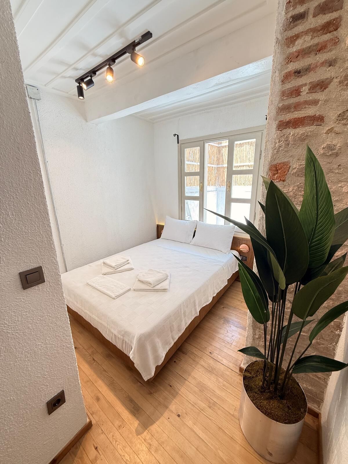 A bedroom with a wooden floor, a bed with white bedding, a potted plant, and a window with a view.