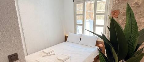 A bedroom with a wooden floor, a bed with white bedding, a potted plant, and a window with a view.