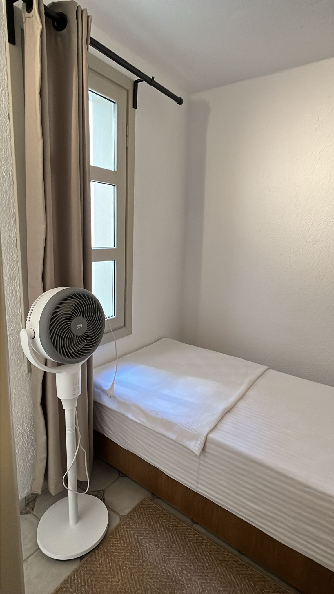 Ein kleines Zimmer mit einem Einzelbett, einem Standventilator, einem Fenster mit Vorhängen und Holzboden.