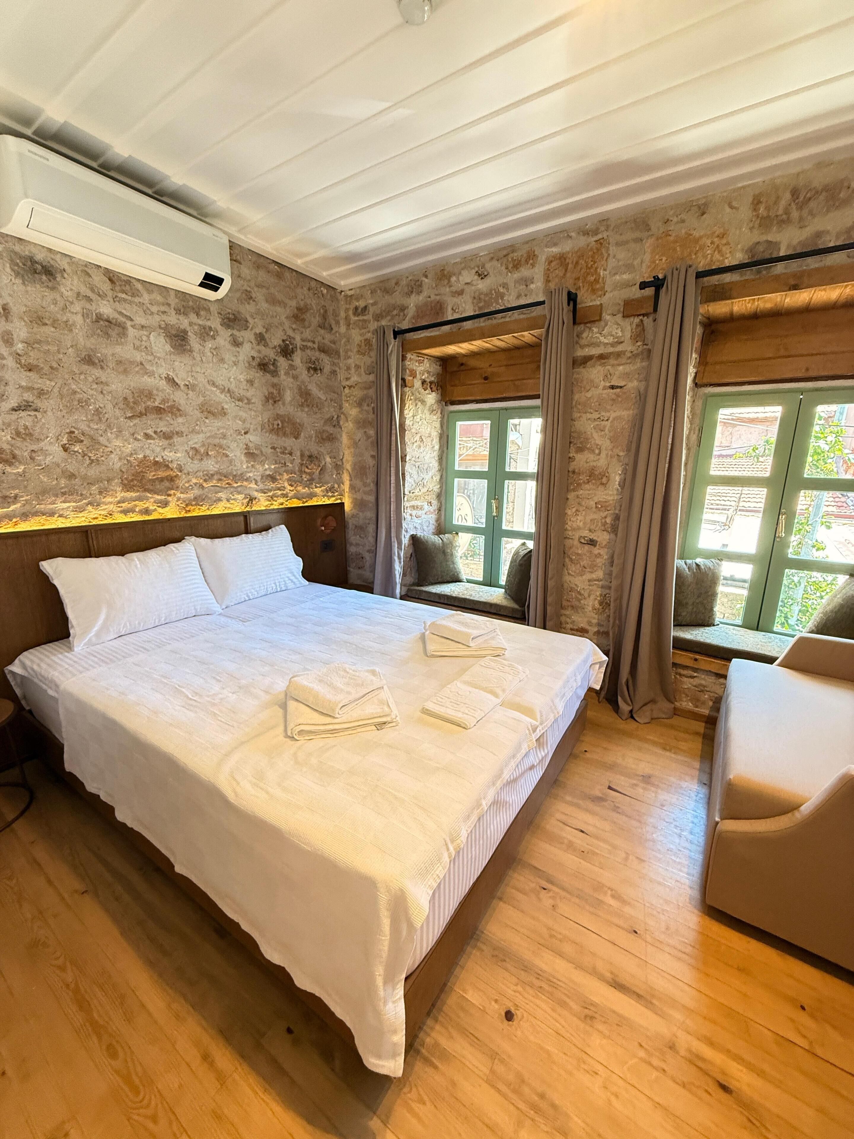 A bedroom with a large bed, wooden flooring, stone walls, a window with curtains, and a built-in bench.