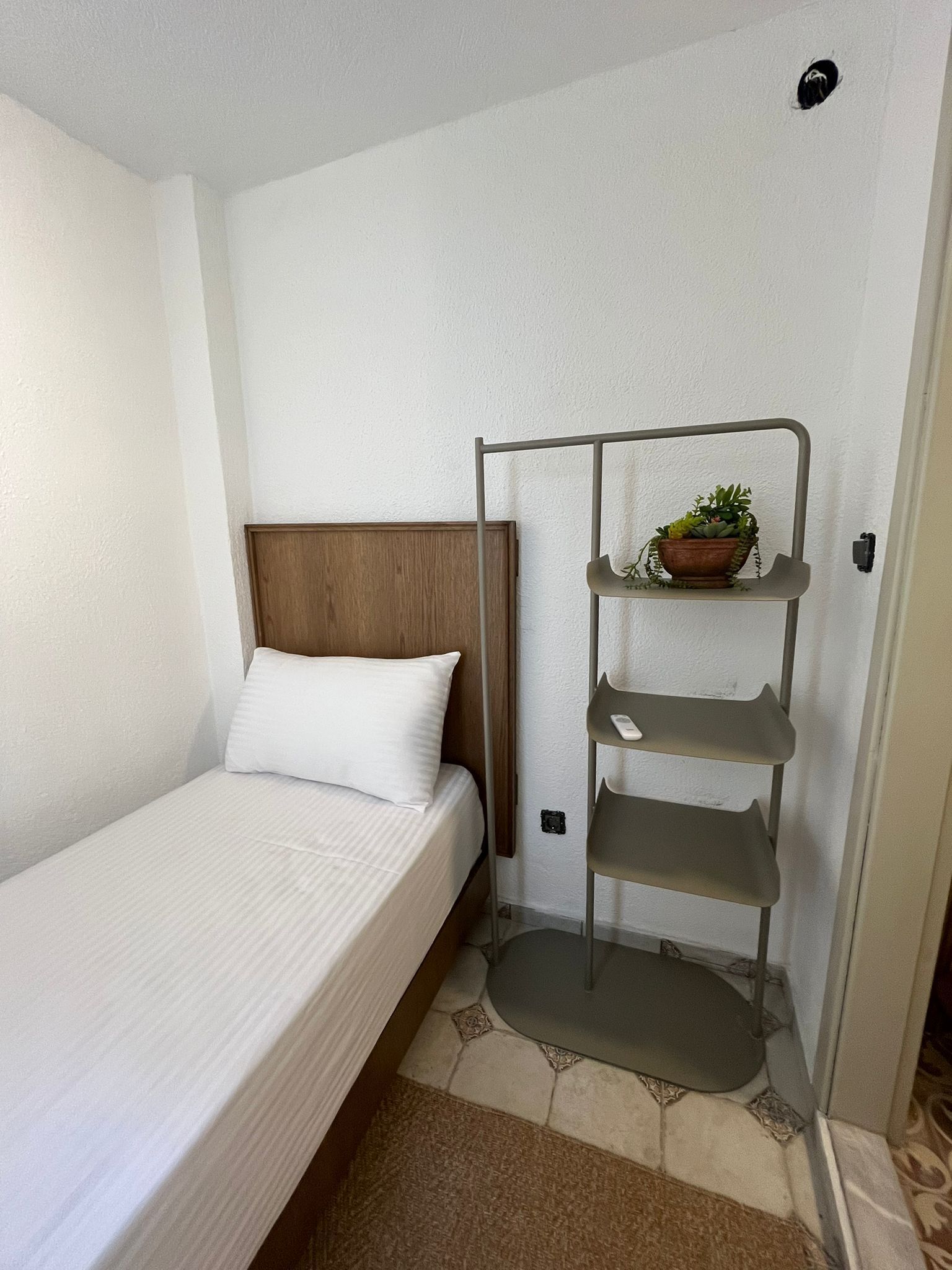 Quarto de hotel pequeno e bem organizado, com uma cama de solteiro, cabeceira de madeira e uma estante de metal com uma planta em vaso.