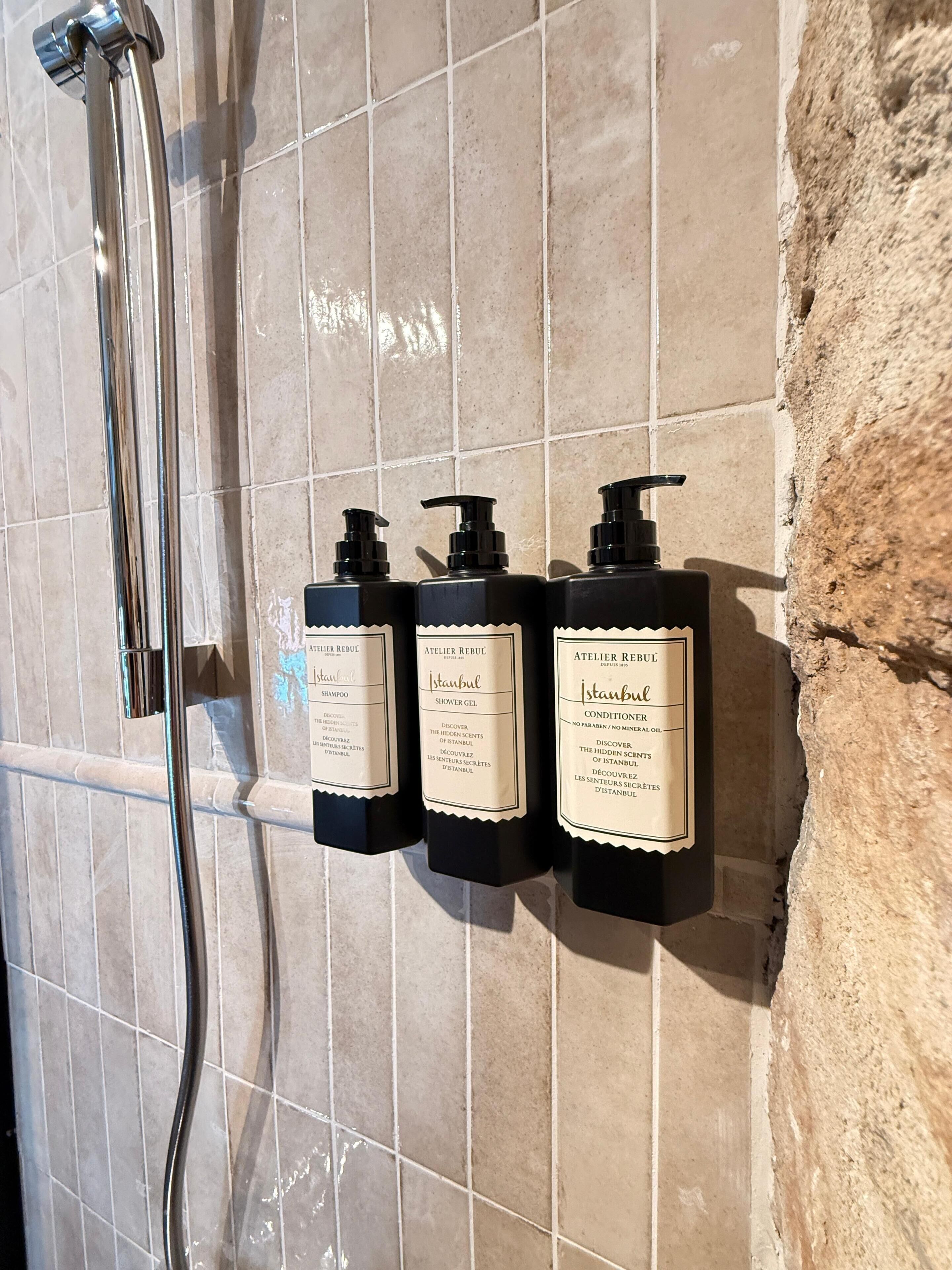 Three black bottles of Istanbul-based shower gel and shampoo on a tiled wall.